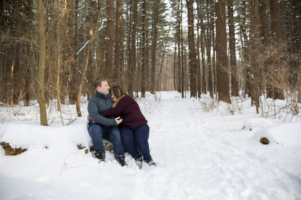 Wife hugs husband while sitting on snow covered log during winter couple's photos