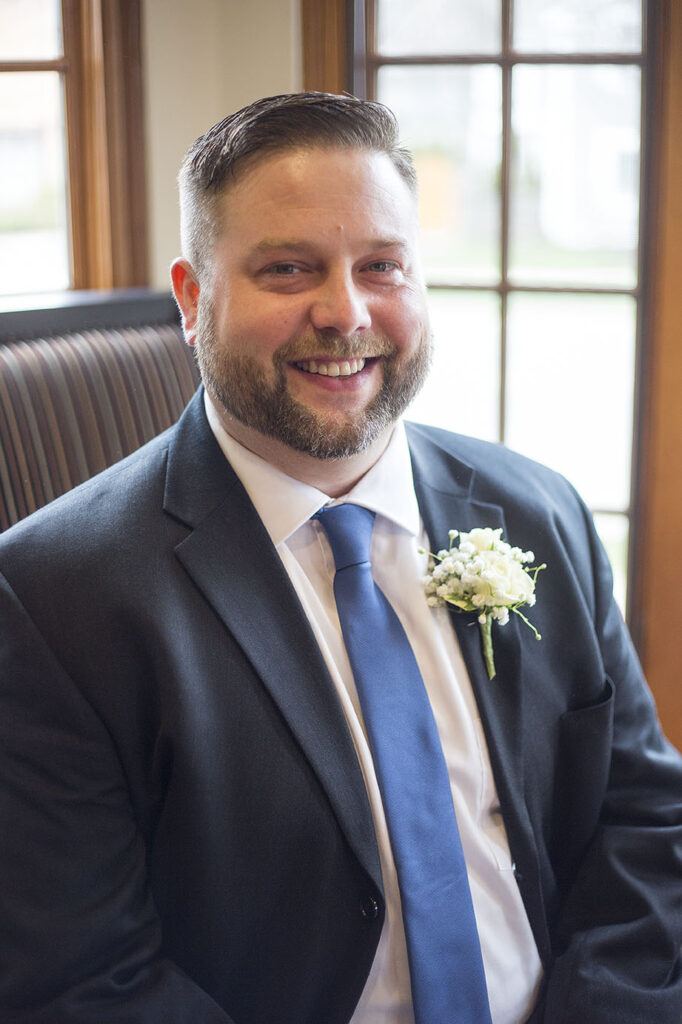 Groom sitting next to large window before his Plymouth wedding