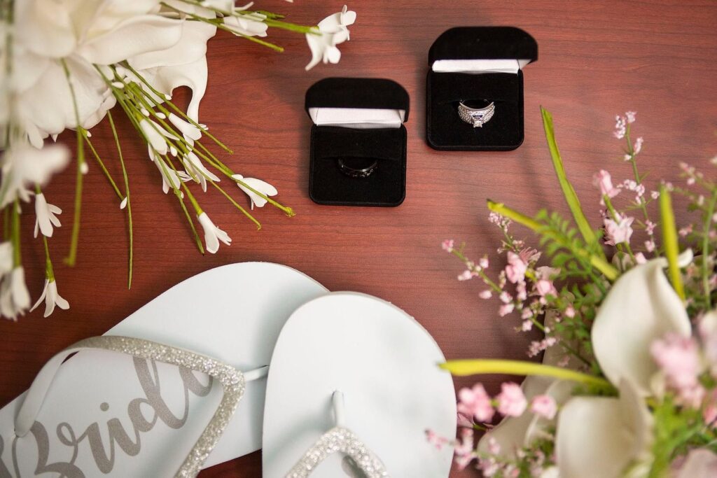 Belleville Michigan wedding photographer arranges "Bride" flip flops, wedding rings, and some florals for a flat lay.