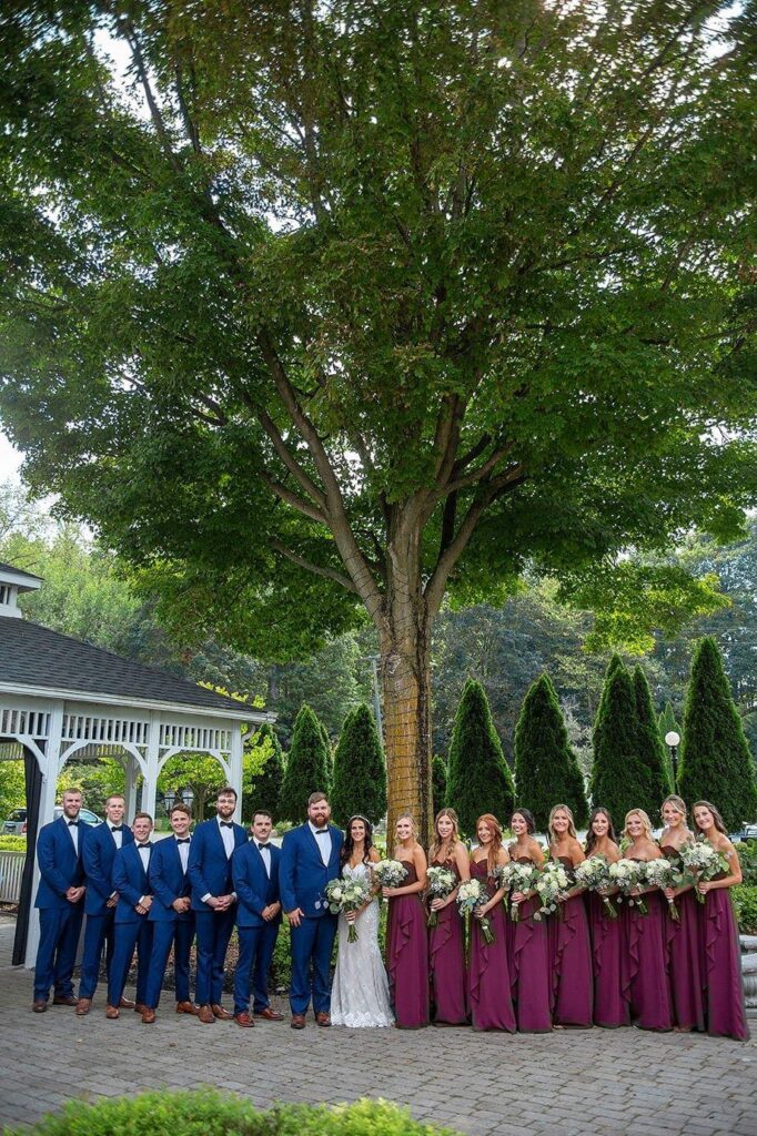 Large wedding party, wearing maroon and navy, gathers beneath a tree outside of Wellers Carriage House.