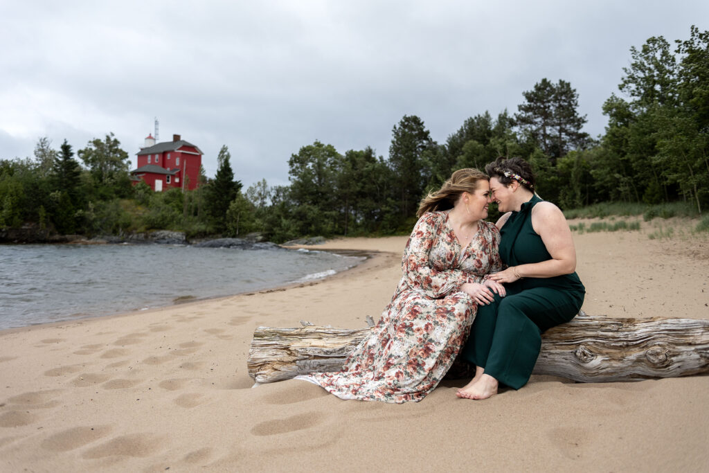 Michigan LGBT wedding couple sit on a driftwood log in front of Marquette Harbor Lighthouse.