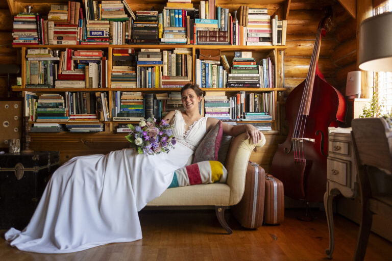 A bride in a flowing white gown lounges gracefully on a chaise lounge, set against a backdrop of a stylish bookcase filled with books and decorative items. The scene is set in Pinecrest Northwoods, Michigan's Upper Peninsula, where the natural beauty of the surroundings complements the elegance of the bride's attire. The serene ambiance highlights the blend of romance and rustic charm characteristic of this unique wedding venue.