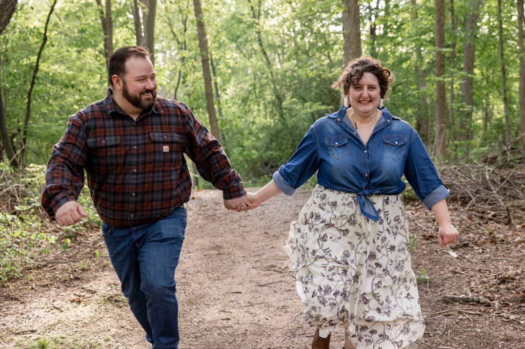 Collin and Natalie running through the woods of Woldumar Nature Center during their engagement session
